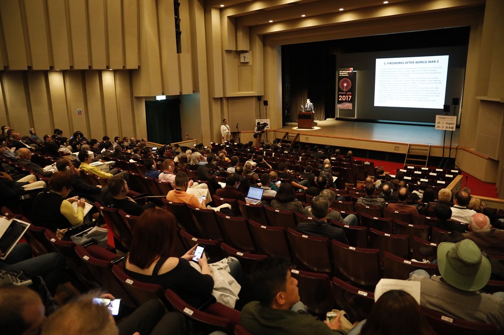 国際花火シンポジウムの様子、ホール内ステージ上にスクリーンを使って説明者が説明している、客席には多くの参加者