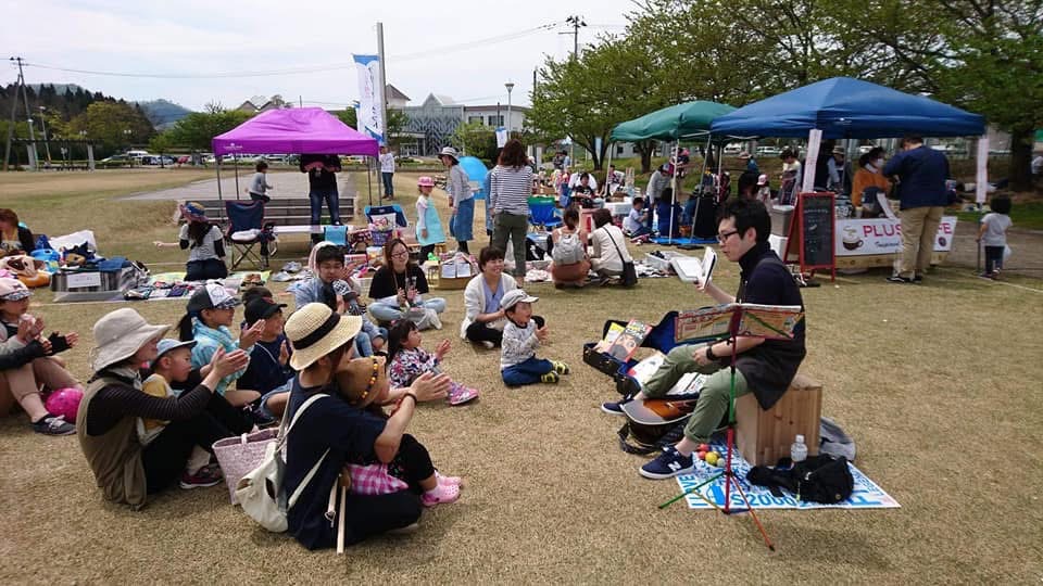 絵本ライブ写真（芝生の上で、男性が読み聞かせしている状況を食いいるように見つめる多くの親子連れ）