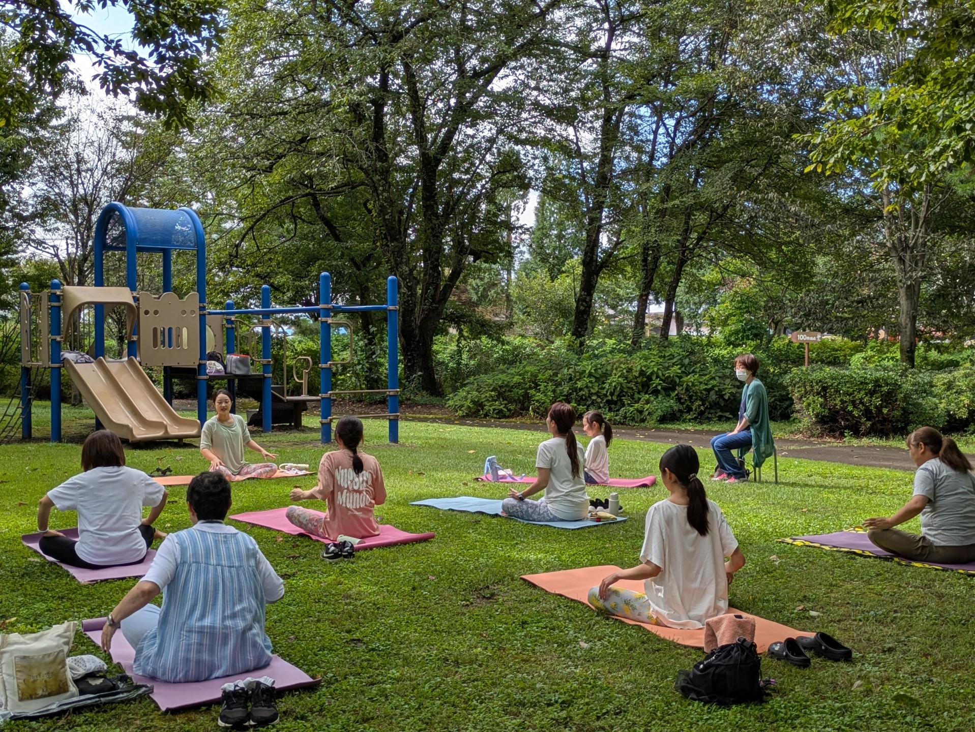 ヨガ教室の写真、青々とした木々に囲まれて木漏れ日のさす芝生の上でヨガを楽しむ参加者たち